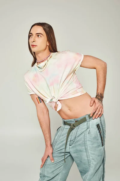 stock image tattooed and young gay man with long hair standing in denim jeans and tied knot on t-shirt showing his belly and posing during pride month on grey background