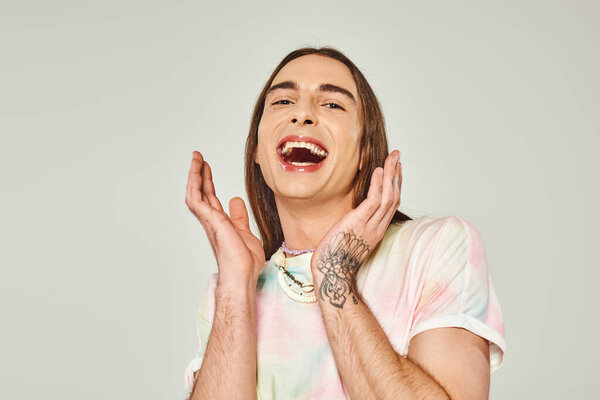 tattooed and excited young gay man with long hair and opened mouth looking at camera and gesturing with hands on grey background, pride month concept