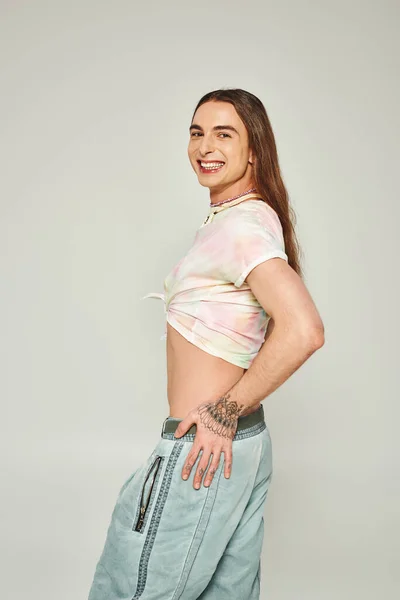 cheerful young gay man with tattoo and long hair standing in denim jeans and tied knot on t-shirt showing his belly and looking at camera on grey background, pride month concept