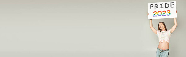 веселый молодой гей-активист с татуировкой и длинными волосами, стоящий в джинсах в джинсах и завязанный узел на футболке, показывающий свой живот и держащий гордость 2023 плакат на сером фоне, баннер 