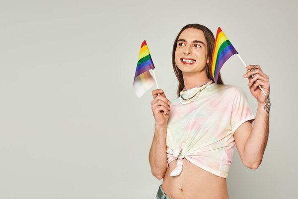 happy and tattooed homosexual man with long hair and bare belly smiling while holding rainbow lgbt flags for pride month on grey background 