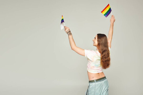 cheerful and tattooed gay man with long hair standing in denim jeans while holding rainbow flags for pride month above head on grey background 