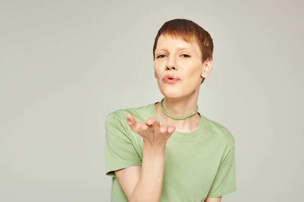 young nonbinary person with lip gloss standing in green t-shirt and blowing air kiss while looking at camera during pride month on grey background