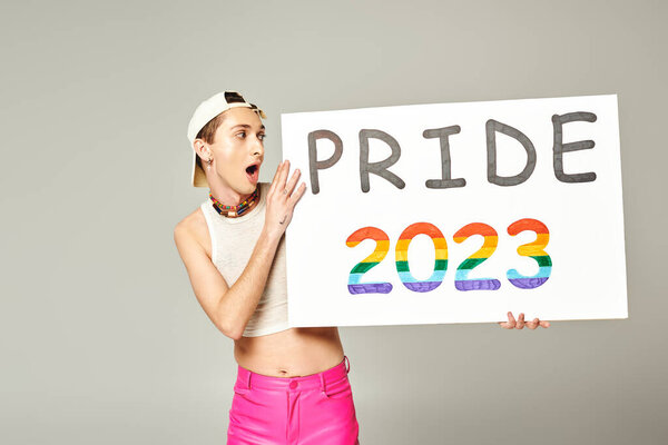 татуированный и шокированный гей в бейсболке, топе и розовых штанах, держащий плакат гордости 2023 года, стоя с открытым ртом на сером фоне 