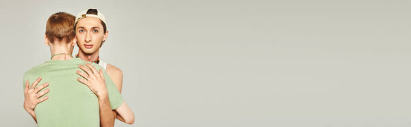 татуированный гей в бейсболке обнимающий и поддерживающий друга сообщества lgbt, глядя в камеру в течение месяца гордости на сером фоне в студии, баннер 