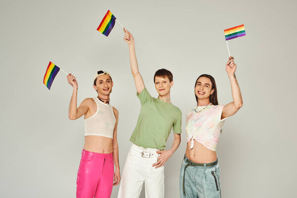 happy and young lgbtq community friends with tattoos standing in colorful clothes and holding rainbow flags while looking at camera in studio, grey background 