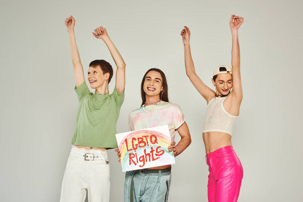 счастливые друзья в красочной одежде танцуют с поднятыми руками рядом с активистом, держащим плакат с надписью lgbtq права и улыбаясь на гордости месяц, серый фон 