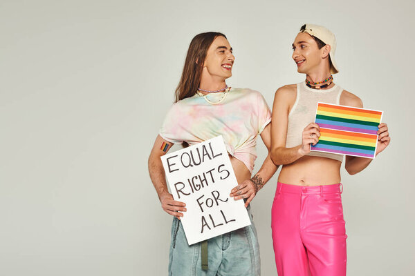 positive and tattooed lgbt people holding rainbow flag picture and placard with equal rights for all lettering while looking at each other on pride day, grey background 