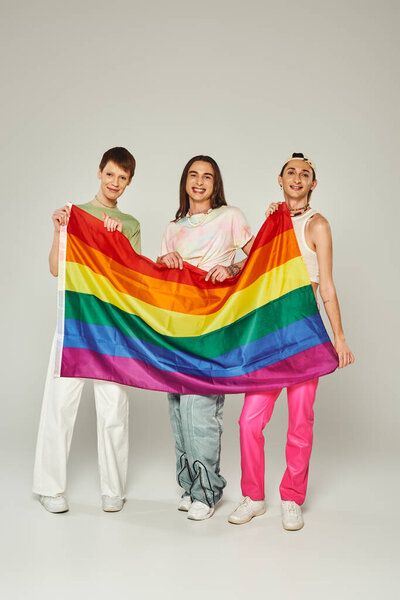diverse group of positive and young friends in colorful clothes holding rainbow lgbt flag and looking at camera while standing together on pride day on grey background