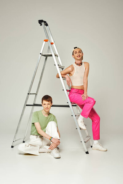 full length of cheerful lgbt friends in colorful and trendy clothes posing next to ladder while smiling together and looking at camera on grey background, pride month concept 
