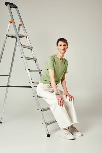 full length of happy queer person with shiny lip gloss posing in green t-shirt and white denim jeans while sitting on ladder during pride month on grey background