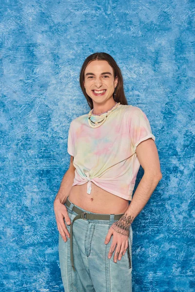 cheerful young gay man with tattoo and long hair standing in denim jeans and tied knot on t-shirt showing his belly during pride month on mottled blue background