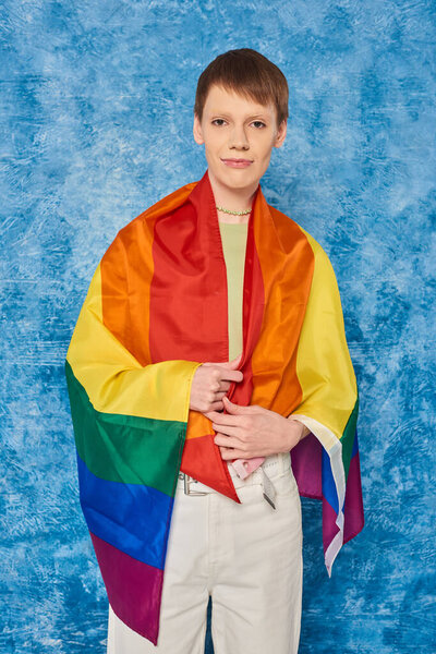 Portrait of young queer person wrapped in lgbt flag looking at camera and standing during pride month celebration on mottled blue background