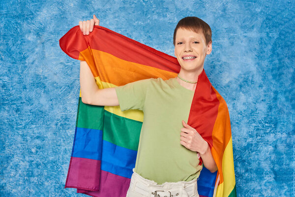 Cheerful gay man in casual clothes smiling and holding lgbt flag and looking at camera during pride month celebration on mottled blue background
