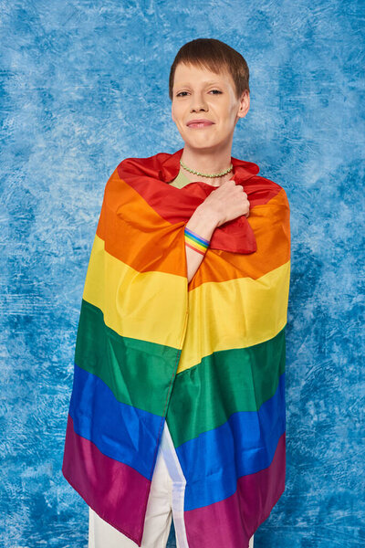 Портрет веселого педика, улыбающегося, держа флаг lgbt и смотрящего в камеру во время празднования месяца гей-прайда на пестром синем фоне