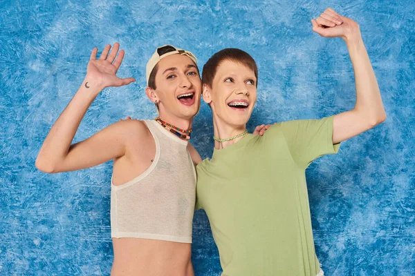 Excited queer people in casual clothes waving hand and hugging during lgbt pride month celebration on textured and mottled blue background