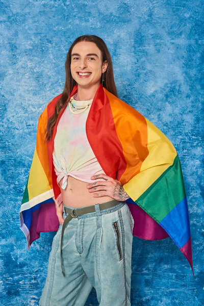 Happy long haired and tattooed gay man in casual clothes with lgbt flag posing and looking at camera while celebrating pride month on mottled blue background