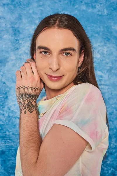 Portrait of young long haired and tattooed gay man looking at camera during lgbt community and pride month celebration on mottled blue background