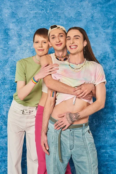 Cheerful homosexual community in stylish outfits hugging each other and looking at camera while celebrating lgbt pride month on mottled blue background