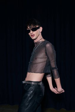 Moda ve güneş gözlüklü genç homoseksüel adam, güneş gözlüklü, kıvılcımlar saçan, deri pantolonlu, Igbt gurur aylarında poz veren, siyahlar içinde izole edilmiş. 