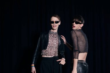 Trendy and young homosexual friends in sunglasses and party outfits posing during lgbt pride month celebration isolated on black  clipart