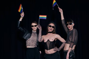 Carefree and fashionable homosexual friends in sunglasses and party clothes holding rainbow flags during pride month celebration isolated on black  clipart