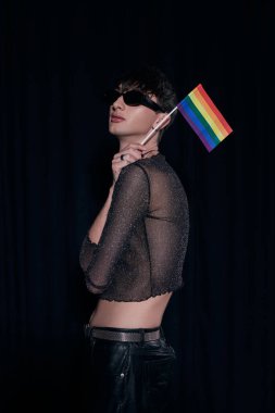 Moda ve güneş gözlüklü genç homoseksüel adam ve parlak tepeli gökkuşağı bayrağı, Igbt gurur aylarında siyahlara izole edilmiş bir şekilde kutlanıyor. 