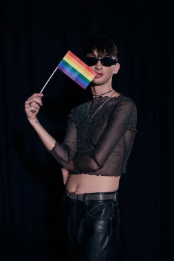 Modayı takip eden güneş gözlüklü genç adam ve parlak parlak üstlü lgbt bayrağı ve gurur aylarında siyahlar üzerinde izole edilmiş bir şekilde poz veriyor. 