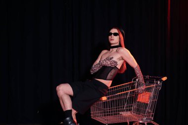 Fashionable homosexual man in sunglasses and sexy corset sitting on shopping cart during party and lgbt pride month celebration on black background  clipart