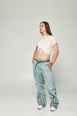 Full length of tattooed and long haired homosexual man holding hands in pockets of denim jeans and looking away during lgbt community pride celebration isolated on grey   clipart