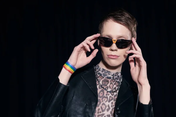 Stylish homosexual man in bracelet with lgbt flag touching sunglasses while posing during pride month celebration party isolated on black