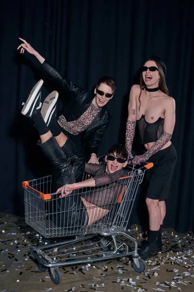 stock image Positive and stylish homosexual community in sunglasses and party clothes having fun with friend in shopping cart on confetti during pride month celebration on black background 