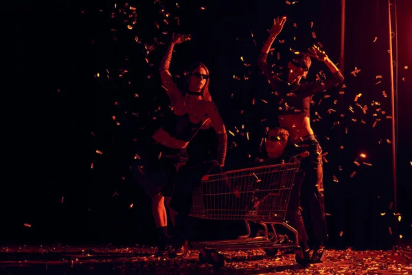 Trendy queer people in sunglasses and party clothes standing near friend in shopping cart under falling confetti during party and lgbt pride month celebration on black background with red lighting