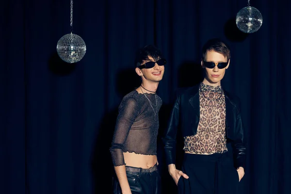 stock image Smiling and fashionable gay friends in sunglasses and party outfits standing near disco balls during lgbt pride month celebration on black background 