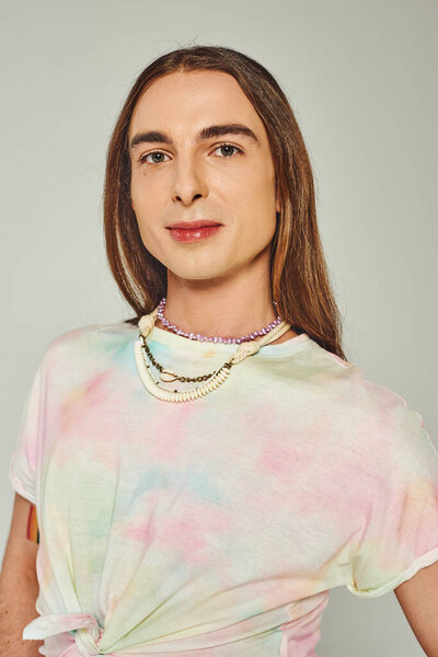 Portrait of young and long haired homosexual man in tie dye t-shirt looking at camera isolated on grey, happy pride community month celebration concept 