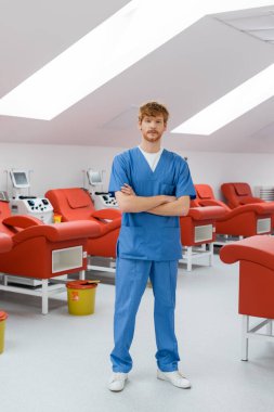 Tam boy kızıl saçlı ve mavi üniformalı genç bir doktor. Kollarını kavuşturmuş, rahat tıbbi sandalyelerin yanında duruyor. Kan bağışı merkezinde otomatik kan nakli makineleri ve çöp kovası var.