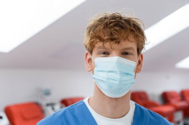 Tıbbi maske takmış genç ve kızıl saçlı bir doktor kan bağışı merkezinde arka planda bulanık duran rahat koltukların yanındaki kameraya bakıyor.