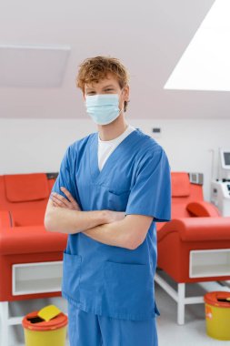 Tıbbi maskeli ve mavi üniformalı genç bir doktor kollarını kavuşturmuş, ergonomik tasarımı olan tıbbi sandalyelerin yanındaki kameraya bakıyor ve kan bağışı merkezinde çöp kovalarına bakıyor.
