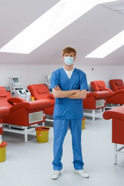 Mavi üniformalı ve tıbbi maskeli kızıl saçlı bir doktor modern hastanedeki tıbbi koltukların, nakil makinelerinin ve çöp kovalarının yanında duruyor.
