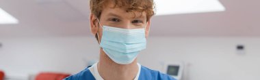 Genç ve kızıl saçlı doktor, neşeli bakışlı, tıbbi maske takan ve steril ortamda kameraya bakan bulanık arka planda kan bağışı merkezi, afiş