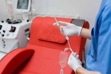 Pratisyen hekimin steril lateks eldivenli kısmi görüntüsü. Tıbbi koltuğun yanında kan nakli ayarlanmış. Rahat ergonomik tasarım ve modern ekipman laboratuvar arka planında bulanık.