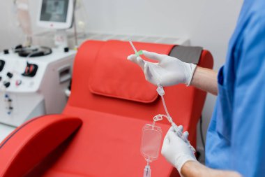 partial view of practitioner in sterile latex gloves holding blood transfusion set near medical chair with comfortable ergonomic design and modern equipment on blurred background in laboratory clipart