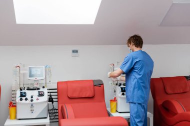 back view of young redhead doctor in blue uniform near automated transfusion machines, plastic cups, drip stands and comfortable medical chairs in blood donation center clipart