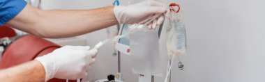 Doktorun lateks eldivenli kısmi görüntüsü. Kan nakli seti ve kan bağışı merkezinin steril ortamında kırmızı tıbbi koltuğun yanında seruma serpiştirme torbaları yerleştiriyor.