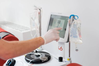 Doktorun steril lateks eldivenli kısmi görüntüsü modern otomatikleştirilmiş transfüzyon makinesi dokunmatik ekranlı serum standının yanında kan bağışı merkezinde infüzyon torbalarıyla