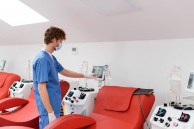 Tıbbi maskeli kızıl saçlı doktor, mavi üniformalı ve lateks eldivenli. Kan bağışı merkezinde, konforlu tıbbi sandalyelerin yanında otomatik kan nakli makinesi var.