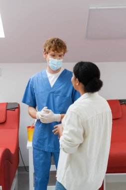 Tıbbi maskeli genç ve kızıl saçlı doktor, lateks eldivenler ve mavi üniformalı kan nakli istasyonunda kırmızı tıbbi sandalyelerin yanında çok ırklı bir hastayla konuşuyor.