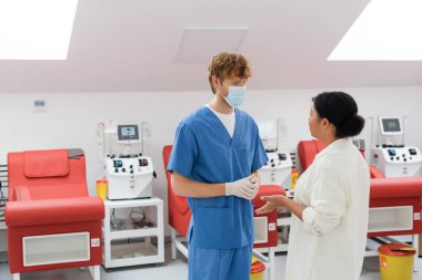 Çok kültürlü bir kadın mavi üniformalı, tıbbi maskeli ve lateks eldivenli genç kızıl saçlı doktorla konuşuyor. Modern laboratuarda kan nakli makineleri ve tıbbi sandalyelerin yanında.
