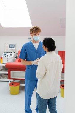 Tıbbi maskeli kızıl saçlı doktor, mavi üniformalı ve lateks eldivenli. Tıbbi sandalyelerin yanında çok ırklı insanlarla konuşuyor ve arka planda bulanık kan nakli makinesi var.