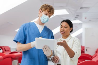 Tıbbi maskeli çok ırklı kadın ve kızıl saçlı doktor, mavi üniformalı ve lateks eldivenler kan nakli istasyonundaki bulanık tıbbi sandalyelerin yanındaki dijital tableti işaret ediyor.